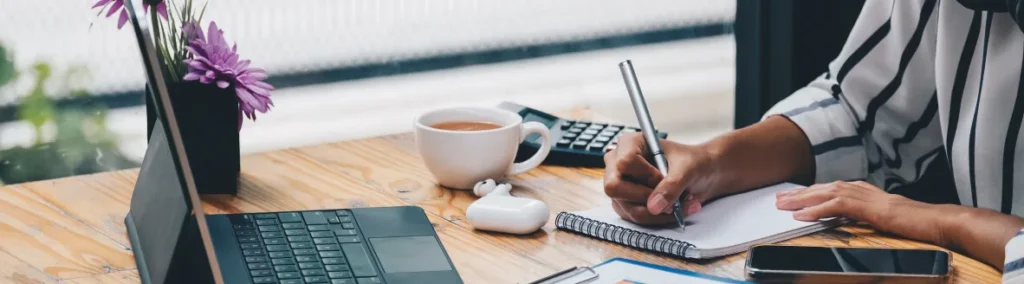 Persona tomando notas en una libreta junto a su portátil y un café